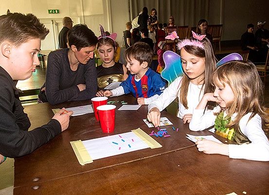 Eine Gruppe von Kindern und Erwachsenen sitzt um einen Tisch herum und verrichtet eine Aktivität. Einige der Kinder tragen Feenflügel und auf dem Tisch liegen rote Becher und Papiere. Der Hintergrund zeigt einen Saal mit stehenden Personen.