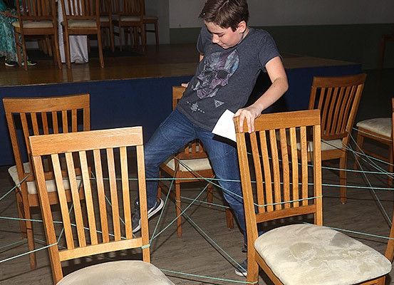 Ein Junge navigiert durch ein Labyrinth aus Stühlen mit grünen Seilen, während er ein Stück Papier in der Hand hält. Er trägt ein graues T-Shirt und blaue Jeans. Die Stühle sind in Reihen auf einem Holzboden angeordnet, und dahinter befindet sich eine Bühne mit einer blauen Plattform.