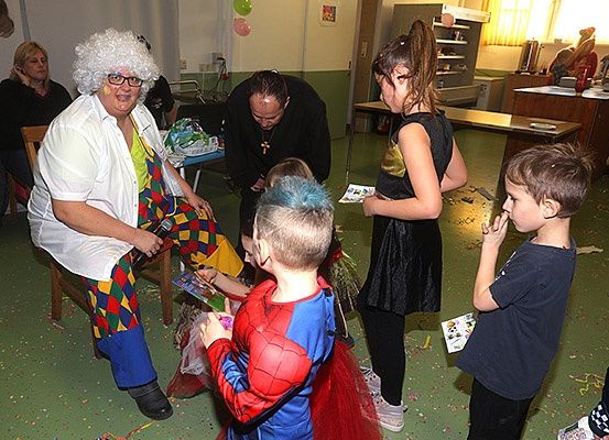 Mehrere Kinder in Kostümen sind um eine Frau in bunter Clown-Kleidung versammelt. Die Frau ist sitzend und hält ein Mikrofon. Andere Erwachsene sind anwesend, einige sitzend und andere stehend im Hintergrund. Es gibt Luftballons und Konfetti auf dem Boden.