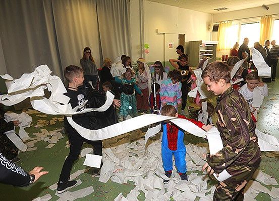 Eine Gruppe von Kindern in Kostümen spielt in einem Raum mit Papier, einige halten Toilettenpapier und andere lachen und schauen zu.