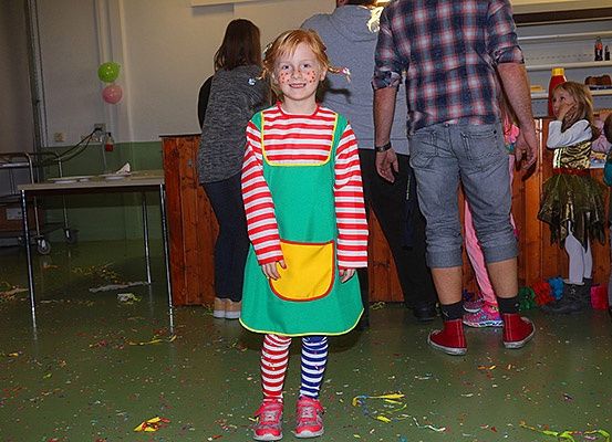 Ein kleines Mädchen in einem grün-weiß gestreiften Kleid steht in einem Raum mit Konfetti auf dem Boden, und mehrere Menschen stehen hinter ihr.