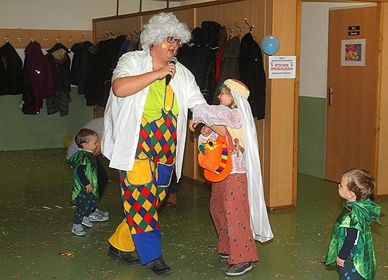Eine als Clown verkleidete Person hält ein Mikrofon und interagiert mit Kindern in Kostümen in einem Raum mit Jacken an der Wand und einer Tür.