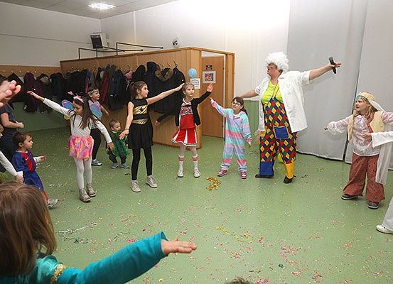 Eine Gruppe von Kindern in Kostümen, einige halten Luftballons, nehmen an einem Spiel oder einer Aufführung teil, mit einer Person in Clownskostüm in einem grünen Raum.