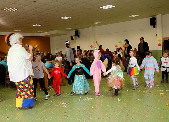 Eine Gruppe von Kindern in Kostümen tanzt in einem Raum mit einem Clown, der ein Mikrofon hält. Sie sind von anderen Menschen umgeben, einige stehen, und der Boden ist mit Konfetti bedeckt.