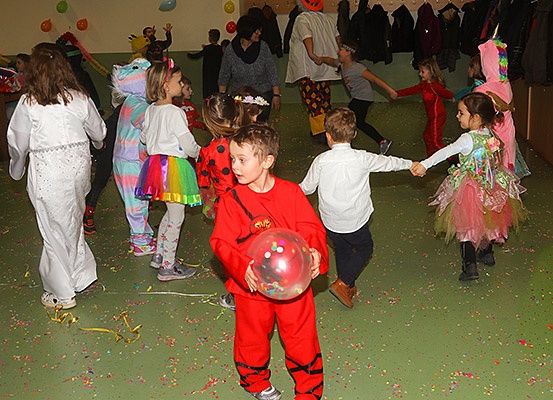 Kinder in Kostümen tanzen in einem Raum mit grünem Konfetti auf dem Boden. Ein Junge in einem roten Kostüm hält einen bunten Ballon.
