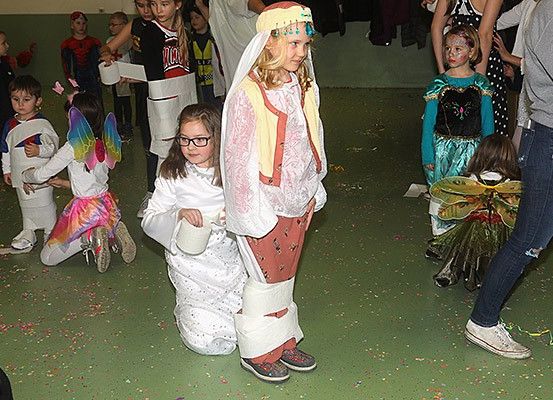Kinder in Kostümen sind drinnen, wahrscheinlich auf einer Halloween-Party. Ein Mädchen in einem weißen Kleid hält einen Becher. Ein Mädchen mit Brille hält ein Kind. Andere sind als Superhelden und Feen gekleidet.