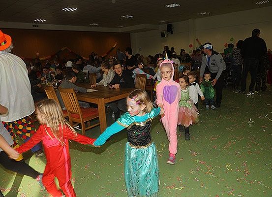 Ein Saal ist mit Menschen gefüllt, viele in Kostümen gekleidet. Einige Kinder halten Händchen und laufen herum. Einige Leute sitzen auf Stühlen um einen Tisch, andere stehen. Ein Lautsprecher ist an der Wand montiert.