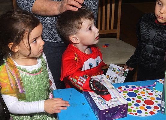 Drei Kinder auf einer Party, eines in einem roten Shirt mit einem Lightning McQueen-Design, ein anderes in einem grünen Kleid und ein drittes in einem schwarzen Kostüm, alle schauen auf eine Geschenkbox auf einem Tisch.