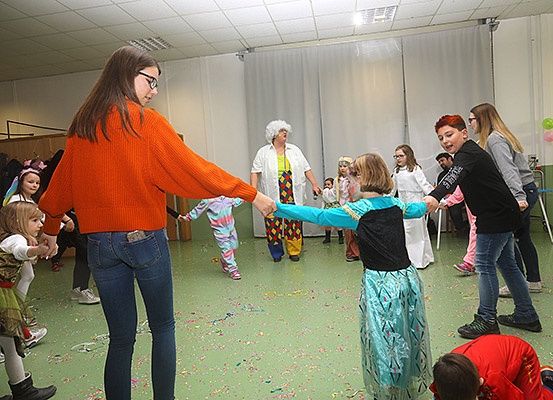 Eine Gruppe von Erwachsenen und Kindern versammelt sich in einem Raum, tanzt und spielt mit Konfetti. Eine Frau in einem orangefarbenen Pullover führt den Tanz an, während sie die Hände eines Kindes in einem blauen Kleid hält. Eine andere Frau in einem weißen Hemd steht hinter ihnen.