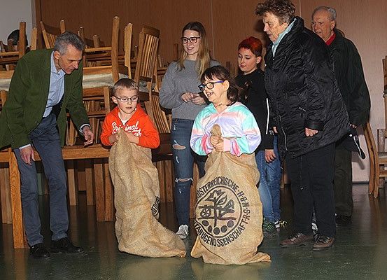 Eine Gruppe von Menschen, darunter Kinder, steht in einem Raum mit hölzernen Stühlen im Hintergrund. Ein Kind hält einen Sack, und andere schauen zu.