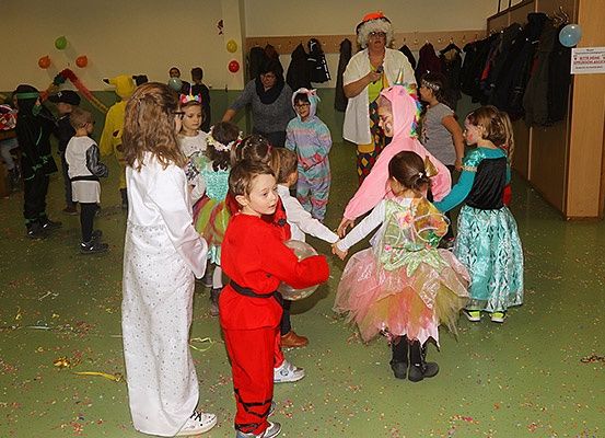 Eine Gruppe von Kindern in Kostümen, wahrscheinlich für eine Party, stehen im Kreis und halten sich an den Händen. Eine Frau in einem weißen Mantel steht vor ihnen, und einige Erwachsene stehen dahinter. Der Raum hat einen grünen Boden und ist mit Luftballons dekoriert.