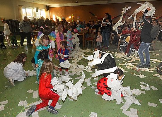 Eine große Gruppe von Kindern und Erwachsenen befindet sich in einem Raum, wahrscheinlich einem Saal, und ist in einer verspielten Aktivität engagiert. Einige halten Stücke Papier, während andere verstreute Stücke aufheben. Der Raum ist mit verstreutem Papier gefüllt. Es gibt Tische und Stühle, und einige Leute stehen in der Nähe der Fenster.