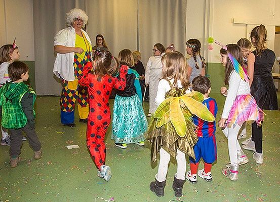 Kinder in verschiedenen Kostümen versammeln sich in einem Raum mit einem Clown. Einige tragen Masken, andere haben Feenflügel. Der Raum ist mit Luftballons dekoriert und hat einen grünen Boden.