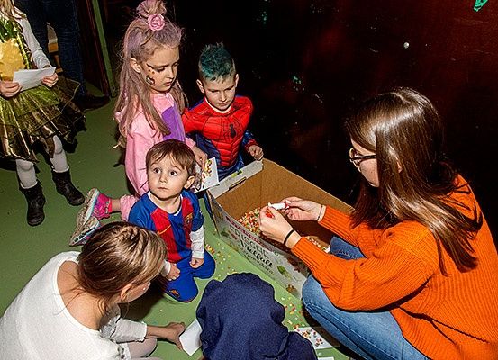 Mehrere Kinder sind um eine Kiste versammelt, möglicherweise an Halloween. Eine Frau in einem orangefarbenen Pullover hält einen Marker und schreibt auf ein Stück Papier. Eine andere Frau in einem weißen Shirt hält ein Papier und sieht die Kinder an. Die Kinder tragen Kostüme.