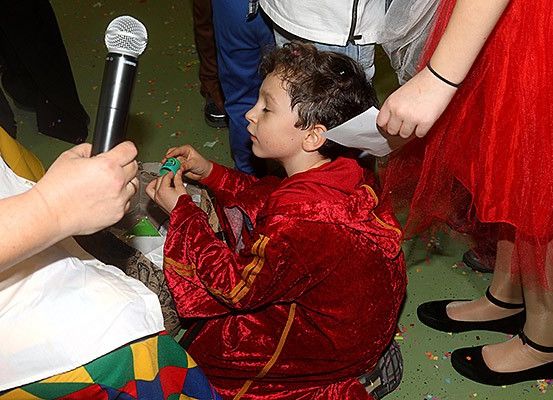 Ein junger Junge in einem roten Kostüm sitzt, hält ein Mikrofon und ein grünes Objekt, möglicherweise ein Spielstein. Ihn umgeben Menschen in verschiedenen Kostümen.