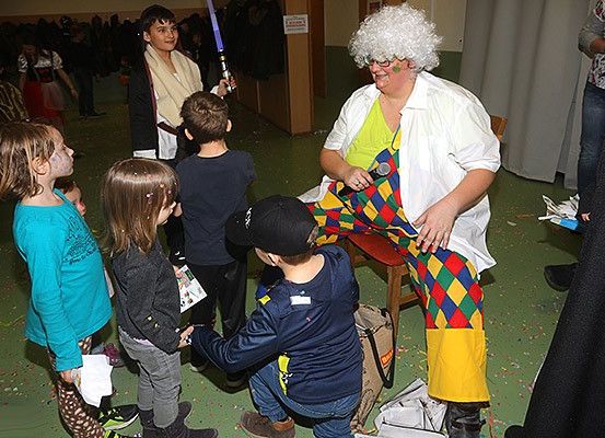 Eine Gruppe von Kindern und Erwachsenen ist um eine Person in einem farbenfrohen Kostüm versammelt, die auf einem Stuhl sitzt. Die Person hält ein Mikrofon und scheint zu sprechen. Die Kinder schauen interessiert zu der Person.