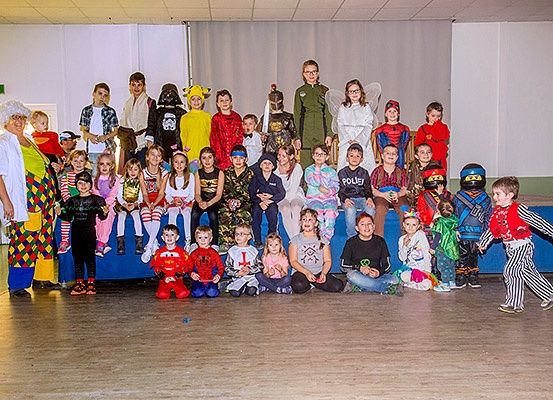 Eine Gruppe von Kindern in verschiedenen Kostümen steht in einem Saal und posiert für ein Foto. Sie stehen auf einer blauen Bank und einem Holzboden.