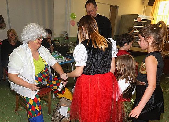 Mehrere Erwachsene und Kinder versammeln sich in einem Raum, wobei eine als Clown verkleidete Person einem Mädchen in einem roten Rock ein Geschenk überreicht. Dahinter steht ein Tisch mit Gegenständen. Ein Mann steht in der Ecke, und es gibt Luftballons und einen Schrank mit einer Mikrowelle oben.