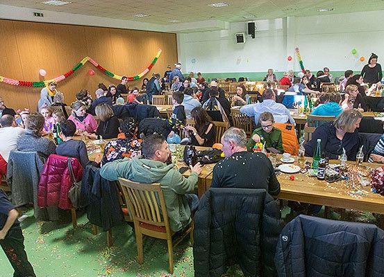 Eine große Gruppe von Menschen sitzt um Tische in einem Saal. Konfetti verteilt sich auf dem Boden. Flaschen, Teller und Tassen stehen auf den Tischen. Einige tragen Jacken und Mützen. Ein Lautsprecher ist an der Wand montiert.