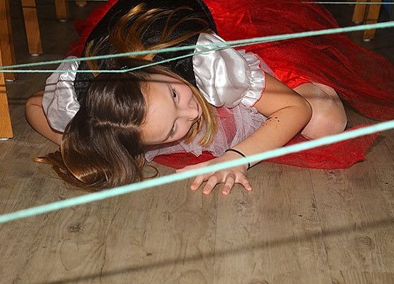 Ein junges Mädchen in einem roten Kleid und einem schwarzen Hut liegt auf dem Boden und navigiert durch ein Labyrinth aus grünen Schnüren. Ihre Haare sind offen, und sie wirkt konzentriert.