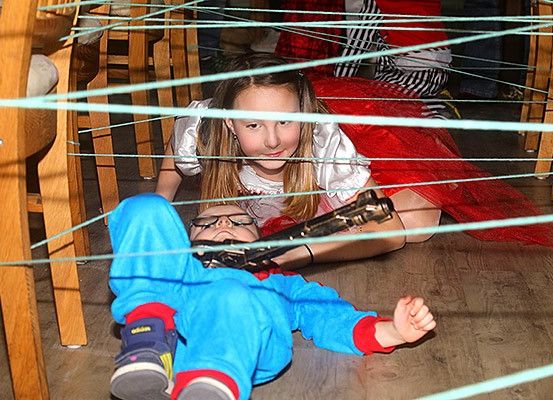 Zwei Kinder spielen in einem mit Schnüren gefüllten Raum, wobei eines auf dem Boden liegt und einen blauen Anzug trägt, während das andere sitzt und eine Spielzeugpistole hält.