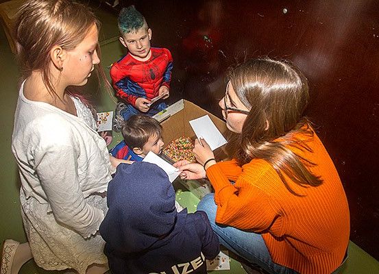 Eine Gruppe von Kindern und zwei Frauen sitzen auf dem Boden. Die Frau auf der rechten Seite hält ein Papier und schaut den Jungen im Spider-Man-Kostüm an.
