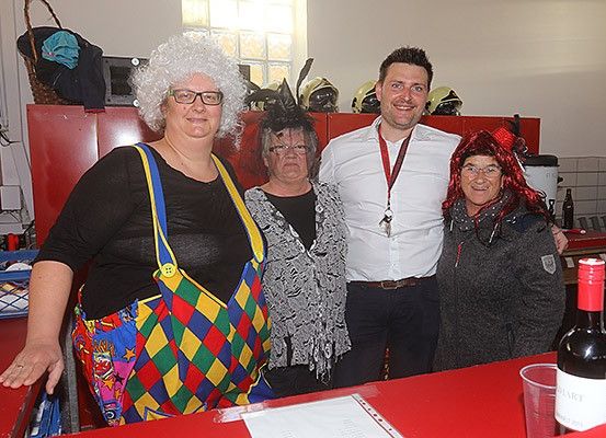 Vier Personen stehen in einem Raum mit einem roten Tresen zusammen, eine Person in Clown-Kleidung und eine andere in einer roten Perücke. Dahinter befinden sich Helme und ein Korb. Flaschen und ein Becher stehen auf dem Tresen.