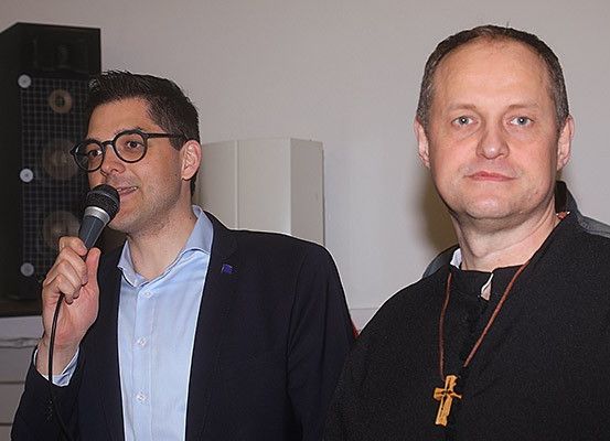 Zwei Männer stehen zusammen; einer hält ein Mikrofon und spricht, der andere trägt eine Kreuzkette. Hinter ihnen befindet sich ein Lautsprecher und eine weiße Lampe.