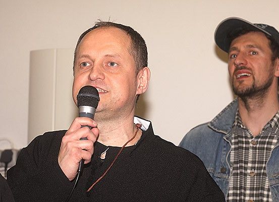 Ein Mann spricht in ein Mikrofon, während ein anderer Mann in einer Jeansjacke und einem karierten Hemd zusieht.