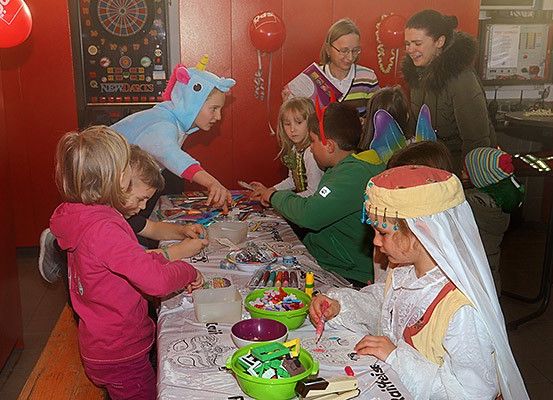 Eine Gruppe von Kindern und Erwachsenen ist an einer Kunstaktivität beteiligt, möglicherweise einem Bastelparty, wobei ein Erwachsener hilft. Die Kinder tragen Kostüme und verwenden Bastelmaterial. Der Tisch ist mit einem weißen Tuch bedeckt und hat verschiedene Gegenstände darauf. Die Wand hinter ihnen hat Dekorationen und ein Dartbrett.