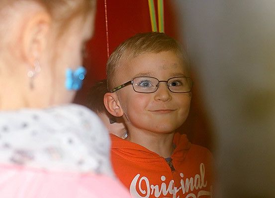 Ein junger Junge mit einer Brille und einem roten T-Shirt mit der Aufschrift 'Original' lächelt, während er neben einer erwachsenen Frau steht.