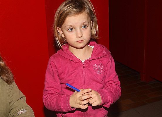 Ein kleines Mädchen mit blonden Haaren hält einen blauen Stift und schaut etwas an. Sie trägt eine rosafarbene Kapuzenjacke mit einem Herzdesign auf der linken Seite. Hinter ihr befindet sich eine rote Wand und eine Tür.
