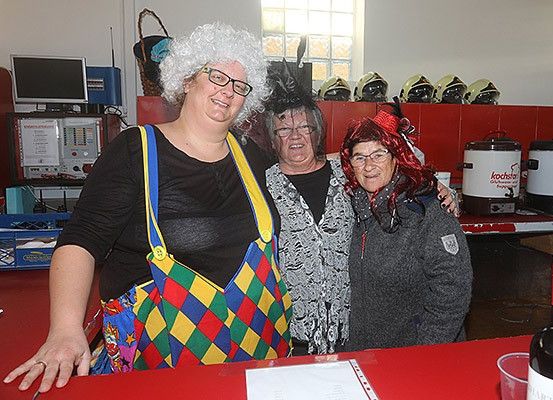 Drei Frauen in Kostümen stehen hinter einem roten Tresen und lächeln für ein Foto. Eine trägt ein Clownkostüm, eine andere einen grauen Pullover und die dritte einen roten Hut. Hinter ihnen sind Helme und eine Kaffeemaschine zu sehen.