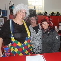 Drei Frauen in Kostümen stehen hinter einem roten Tresen und lächeln für ein Foto. Eine trägt ein Clownkostüm, eine andere einen grauen Pullover und die dritte einen roten Hut. Hinter ihnen sind Helme und eine Kaffeemaschine zu sehen.