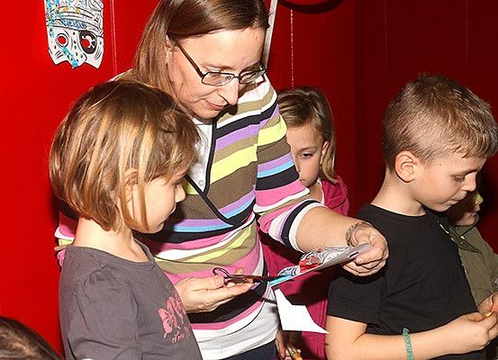 Eine Frau mit Brille hilft zwei Kindern mit Scheren und einem Papierbastelprojekt. Hinter ihnen befindet sich eine rote Wand mit Dekorationen.