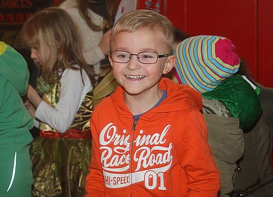 Ein junger Junge mit Brille und einer orangefarbenen Kapuzenjacke lächelt in die Kamera, umgeben von Kindern in festlichen Kostümen.