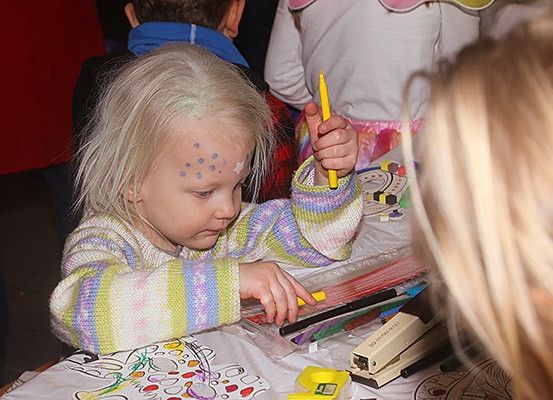 Ein kleines Mädchen mit Gesichtsbemalung zeichnet mit einem gelben Marker auf einem Papier, das auf einem Tisch liegt, eine andere Frau beobachtet.