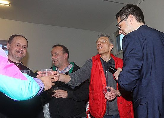 Fünf Männer stehen in einem Raum. Ein Mann in einer roten Weste hebt sein Glas zum Anstoßen. Ein anderer Mann in Anzug und Brille schaut zu.