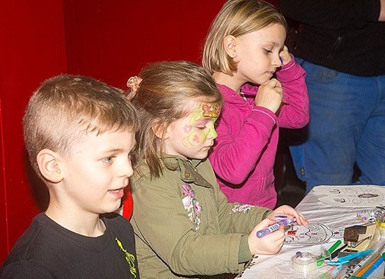 Drei Kinder, zwei Mädchen und ein Junge, sitzen an einem Tisch. Sie malen eine Papierbastelei mit Markern aus. Die Mädchen haben Gesichtsbemalung und eines hält einen Marker. Der Junge hat seinen Kopf zur Seite gedreht. Hinter ihnen ist eine rote Wand.