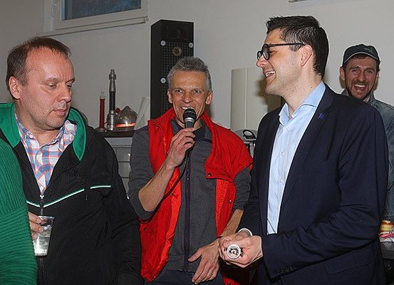 Vier Männer stehen in einem Raum. Ein Mann hält ein Mikrofon und scheint zu sprechen. Die anderen lächeln und lachen. Dahinter sind ein Lautsprecher und eine Lampe zu sehen.