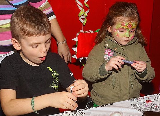 Zwei Kinder, ein Junge und ein Mädchen, malen auf einem Tisch. Der Junge trägt ein schwarzes T-Shirt mit einem Dinosaurier. Das Mädchen hat grünes Gesichtsmal und eine Krone. Hinter ihnen steht eine Person in einem gestreiften Shirt in der Nähe einer roten Wand.