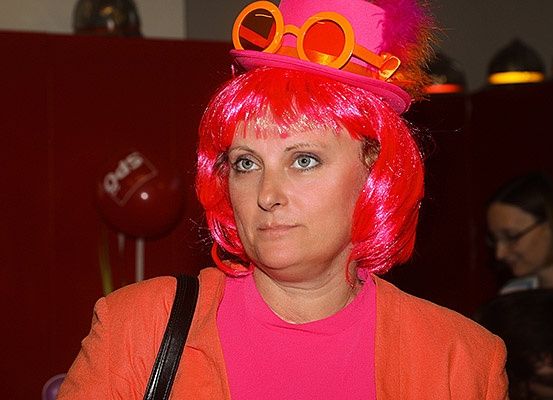 Eine Frau mit rosa Haaren und einem rosa Hut mit orangefarbenen Gläsern. Sie trägt ein rosa Hemd und eine orange Jacke. Hinter ihr befindet sich eine rote Wand mit einem roten Luftballon.