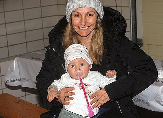Eine lächelnde Frau hält ein Baby in den Armen. Das Baby trägt einen weißen Hut und hat einen Schnuller im Mund. Beide sind in Winterkleidung gekleidet. Hinter ihnen ist eine geflieste Wand mit einer weißen Tischdecke und etwas Text.