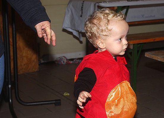 Ein kleines Kind in einem roten und orangefarbenen Kostüm läuft und wird von einem Erwachsenen gehalten. Dahinter befinden sich Holzbänke und Tische.