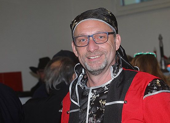 Ein Mann mit Bart trägt ein rotes und schwarzes Outfit und eine Brille. Er lächelt für ein Foto, mit Menschen im Hintergrund.