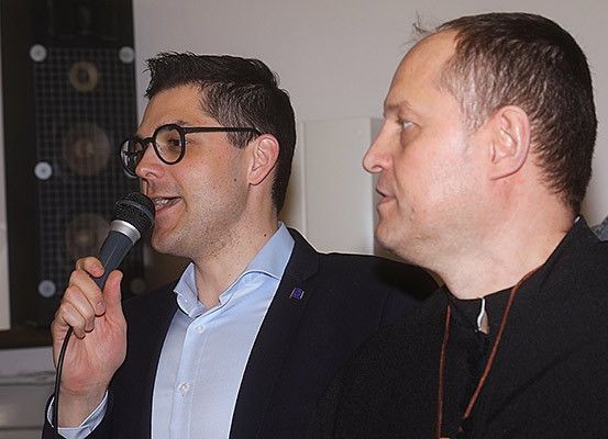 Ein Mann mit Brille spricht in ein Mikrofon, während er einem anderen Mann in einem schwarzen Hemd mit einer Halskette gegenübersteht, vor einem Hintergrund einer weißen Wand.