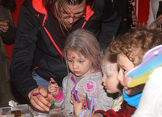 Ein Mann mit Brille hilft einer Gruppe von Kindern mit Gesichtsbemalung, auf einem Tisch zu malen. Die Kinder tragen Jacken.