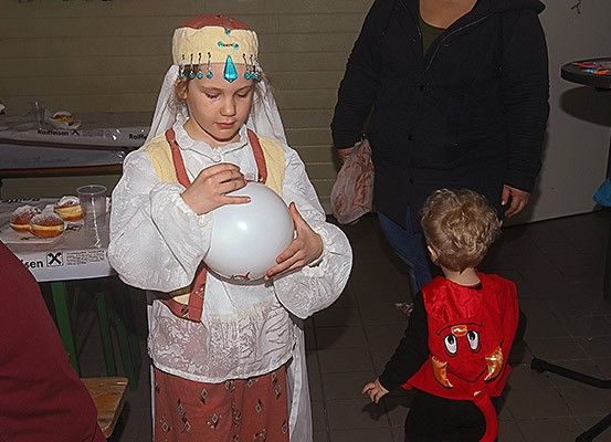 Ein junges Mädchen in traditioneller Kleidung hält einen großen Ballon. Ein Kind in einem roten Kostüm steht in der Nähe. Eine Frau mit einer Handtasche ist im Hintergrund. Ein Tisch mit Gebäck ist ebenfalls zu sehen.