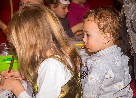 Ein junges Mädchen mit langen Haaren malt, während ein Kleinkind mit zerzausten Haaren sie ansieht. Dahinter hält eine Frau ein Baby. Im Hintergrund beschäftigen sich Kinder mit verschiedenen Aktivitäten, und eines hält einen Stift.