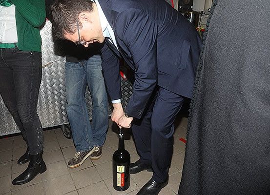 Ein Mann in einem Anzug öffnet eine Weinflasche auf dem Boden, während andere in einem Raum zuschauen.